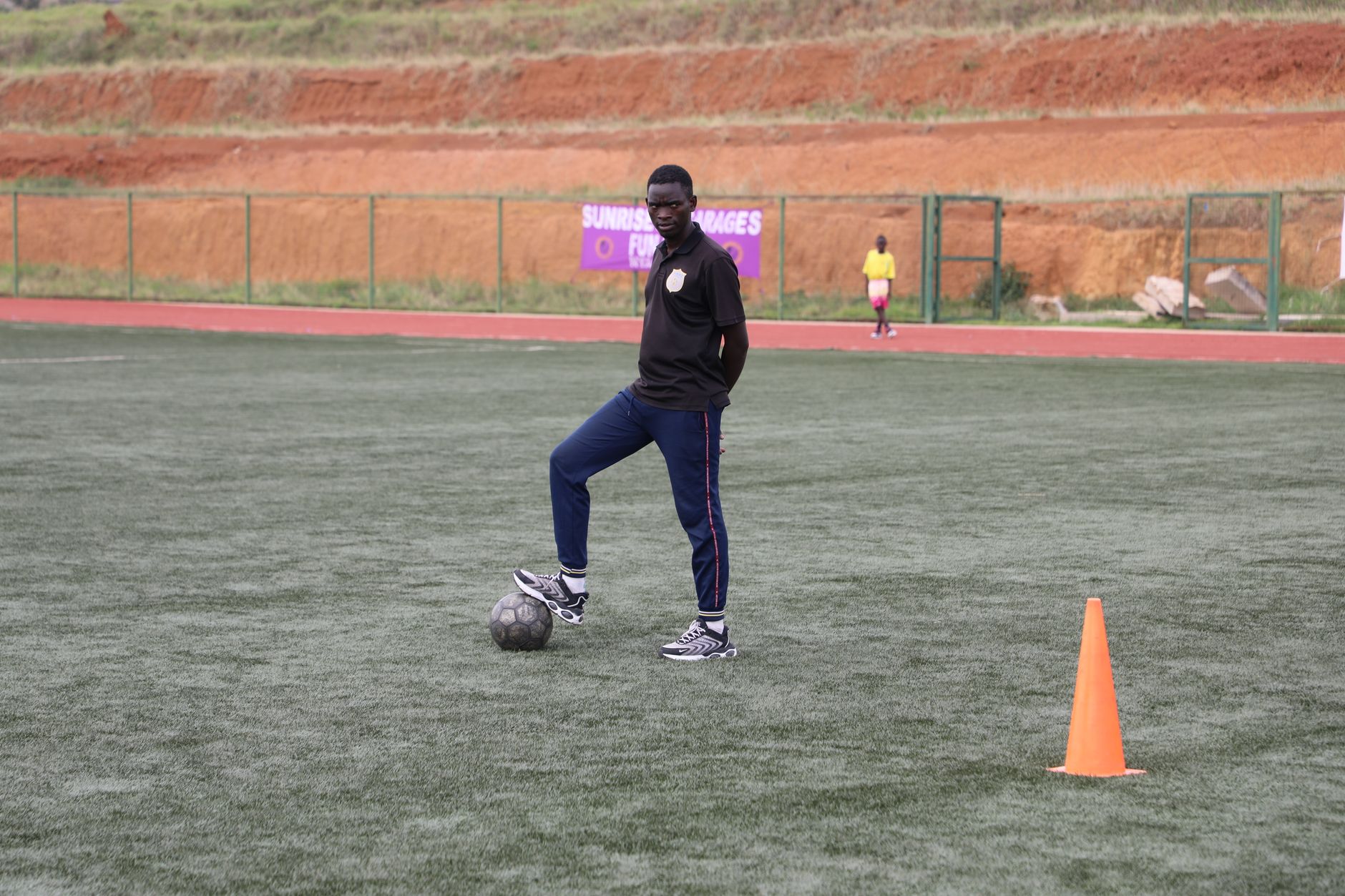 Une personne pose un pied sur un ballon de football, avec un cône orange au premier plan.