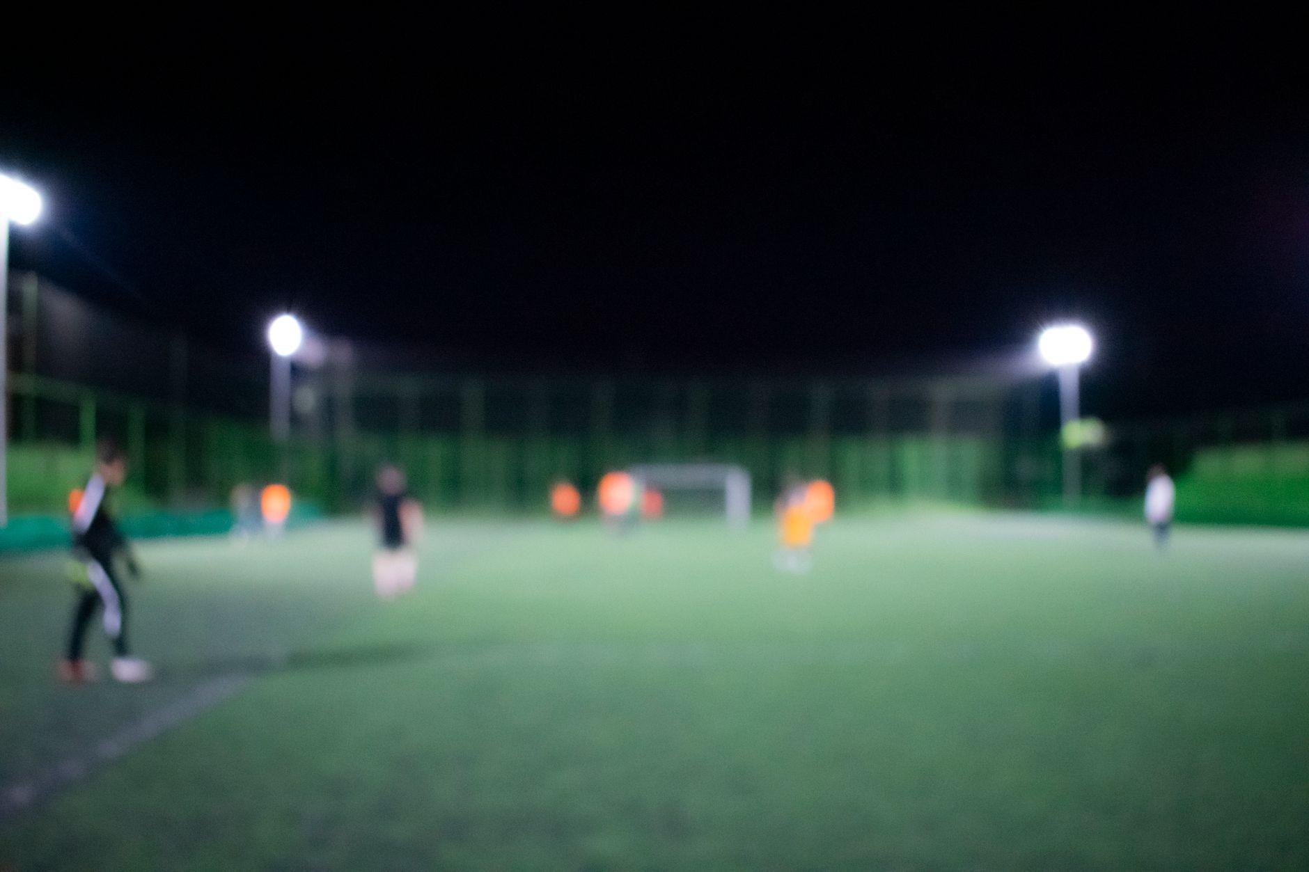 Terrain de football éclairé de nuit par des projecteurs, image floue avec quelques joueurs et un but au fond.